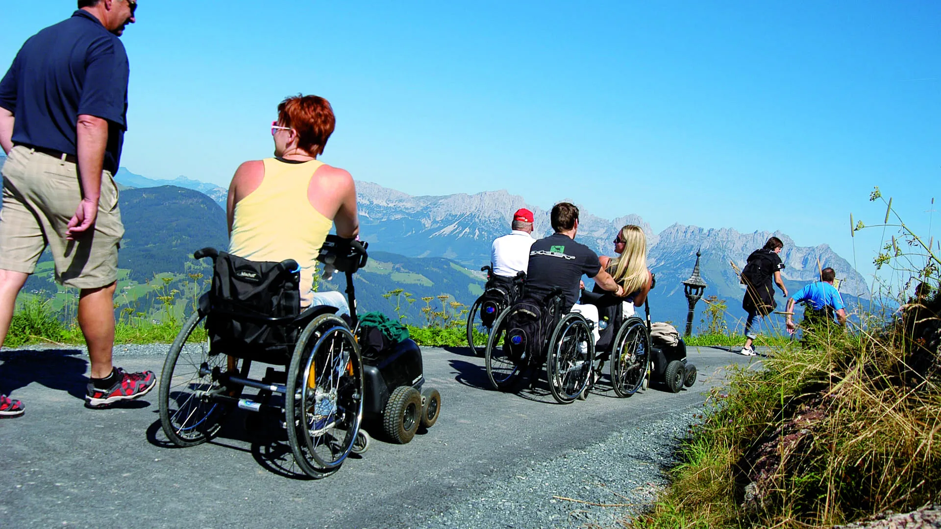 Menschen in Rollstühlen auf einem Weg mit Bergblick, andere gehen vorbei