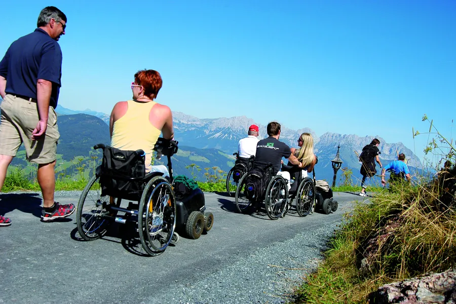 Menschen im Rollstuhl auf einem Weg mit Bergblick, einige gehen zu Fuß.