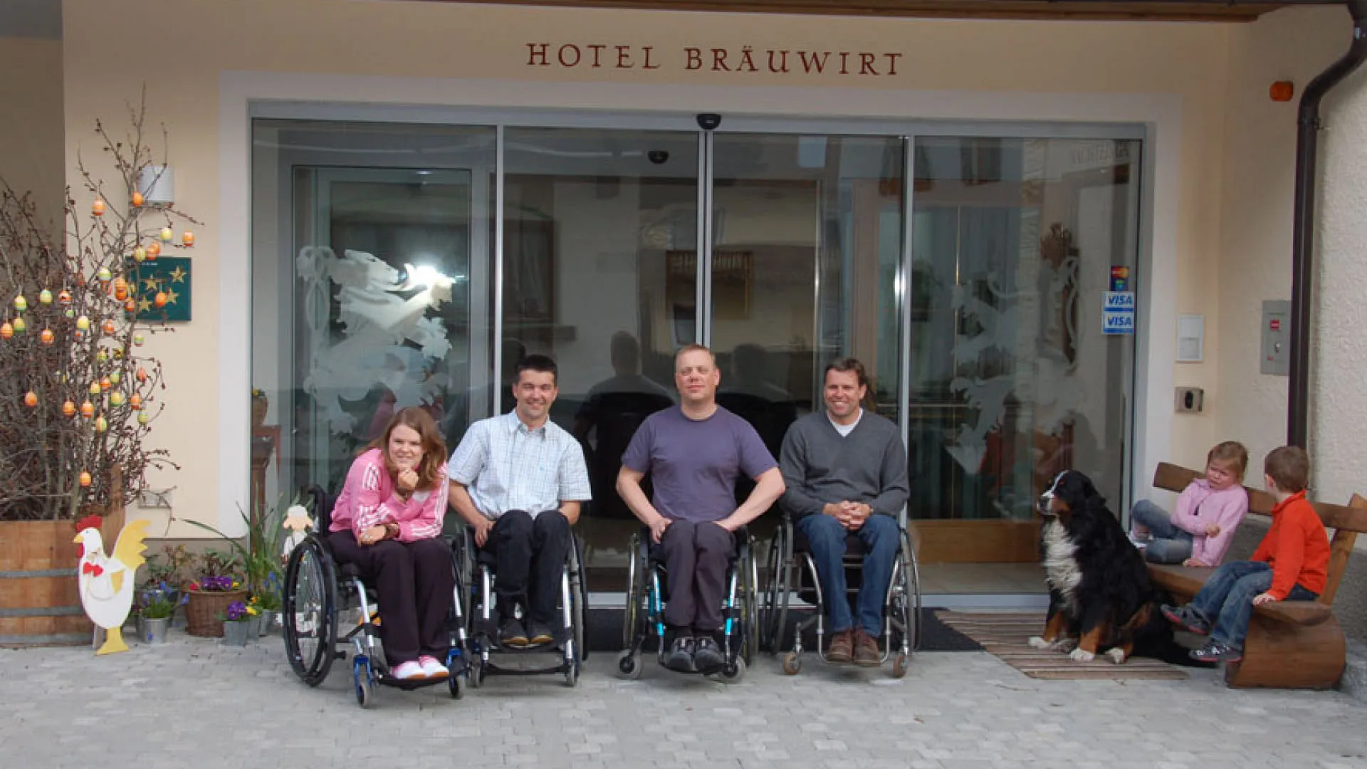 Vier Personen in Rollstühlen vor dem Hotel Brauwirt, Hunde und Kinder im Hintergrund.