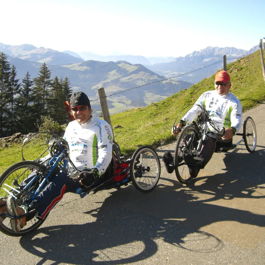 Zwei Männer auf Handbikes fahren eine Straße mit Berglandschaft im Hintergrund entlang