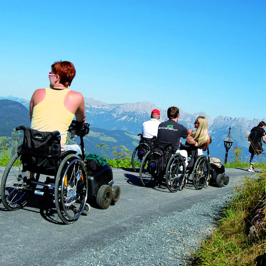 Menschen mit Rollstühlen auf einem Weg mit Bergen im Hintergrund
