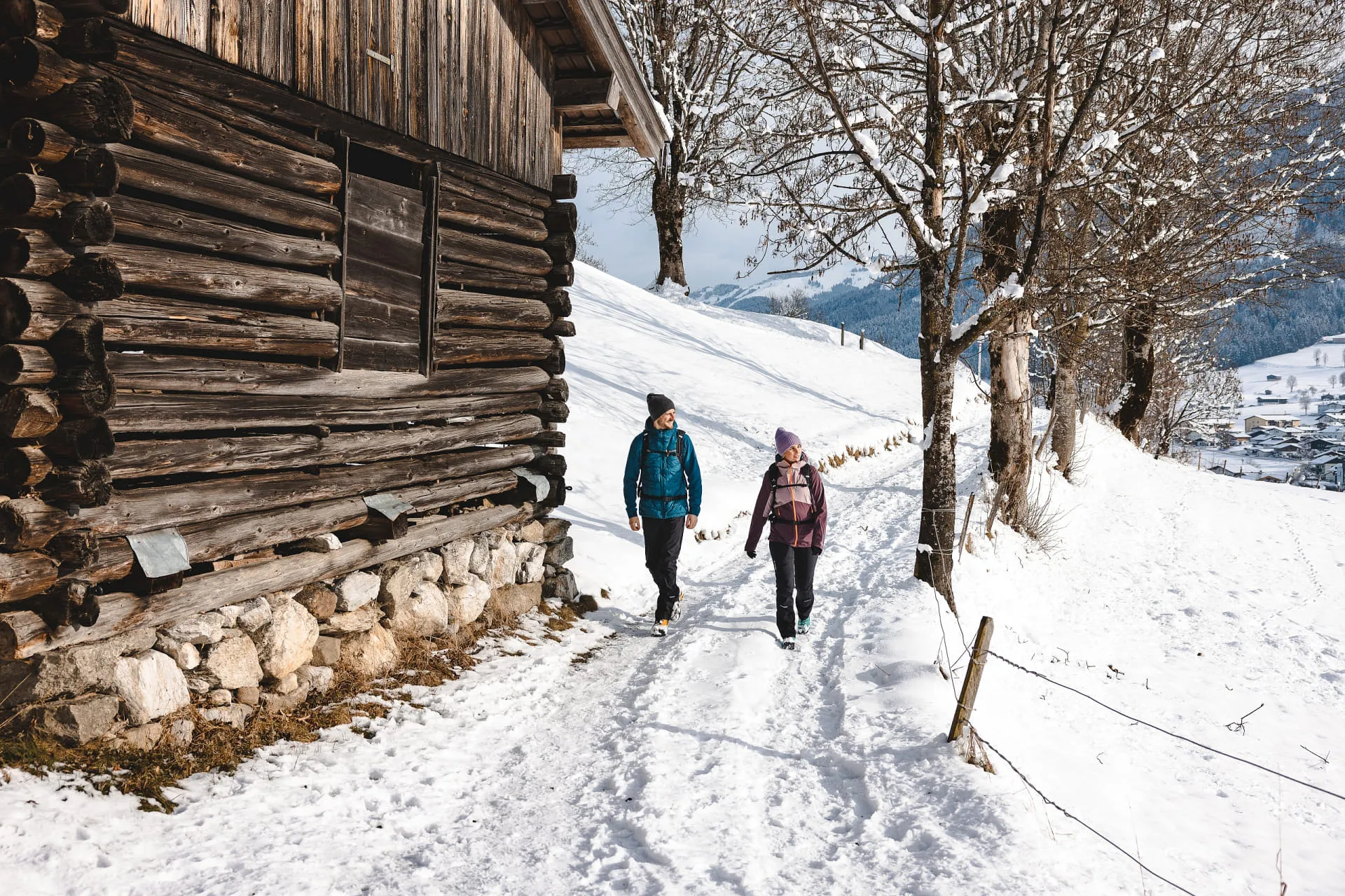 Zwei Wanderer gehen auf einem verschneiten Weg neben einer Hütte im Brixental