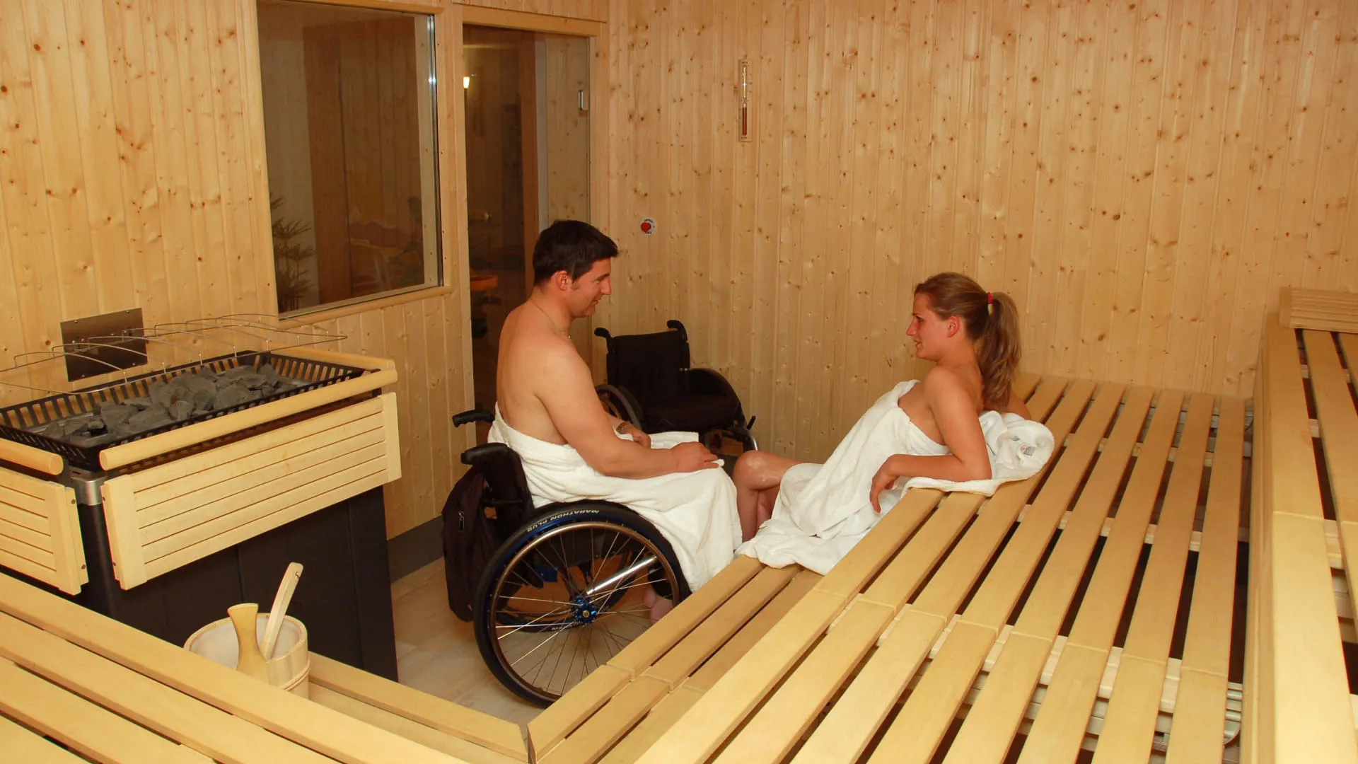 Zwei Personen in einer Sauna; ein Mann im Rollstuhl und eine Frau in einem Handtuch.
