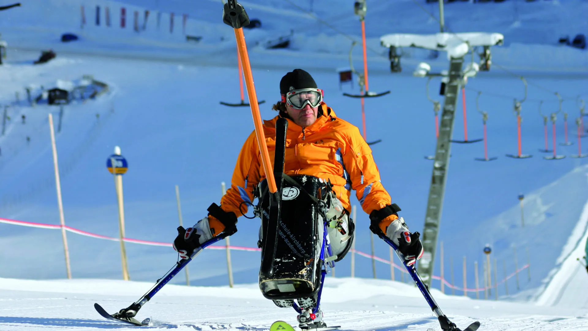 Skifahrer in orangeem Anzug auf einer Piste im Schnee.